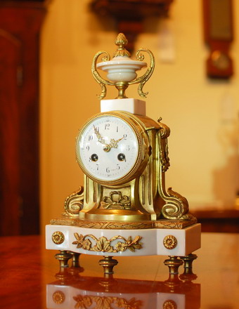A very petite French mantel clock on a white Marble base with Ormalu case and decorations , the clock is of 8 day duration , striking hourly and half hourly on a bell with a white convex enamel dial showing arabic Numerals and finely pierced Gilded Brass Hands , and a cast brass bezel surround . This clock has a very small 
